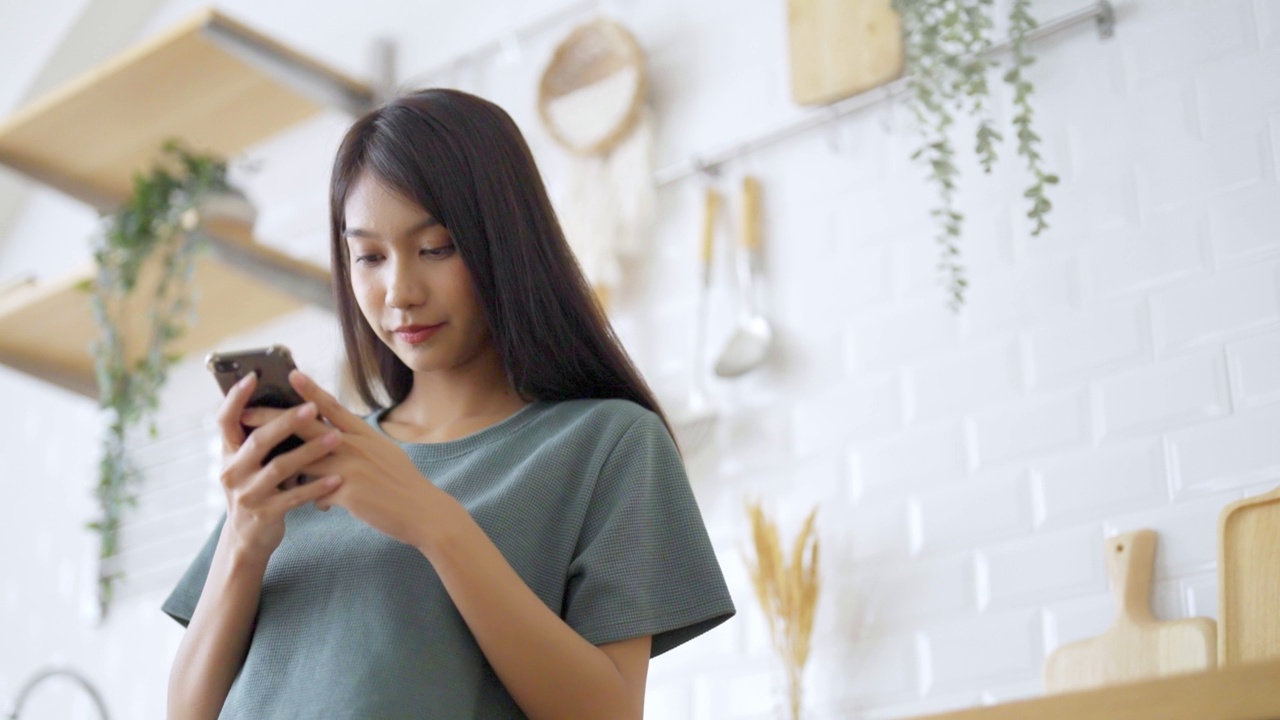 快乐的年轻亚洲女人在家里放松。亚洲女性站在厨房柜台，使用手机视频素材