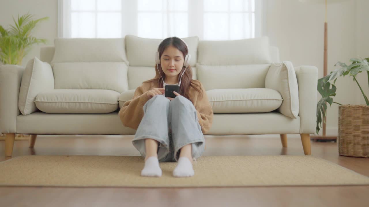 快乐的年轻亚洲女人在家里放松。女性微笑着坐在沙发上，手里拿着手机。女孩用视频电话给朋友视频素材