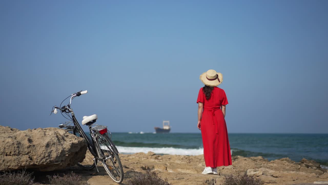 宽镜头美丽的塞浦路斯海景与年轻女子在红色连衣裙欣赏海站在沙滩上的自行车旁边。千禧一代高加索游客在户外享受夏日假期的模板。视频素材