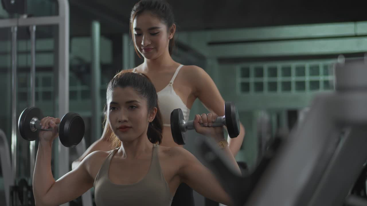 亚洲:年轻女子在健身房里和教练一起举哑铃视频素材