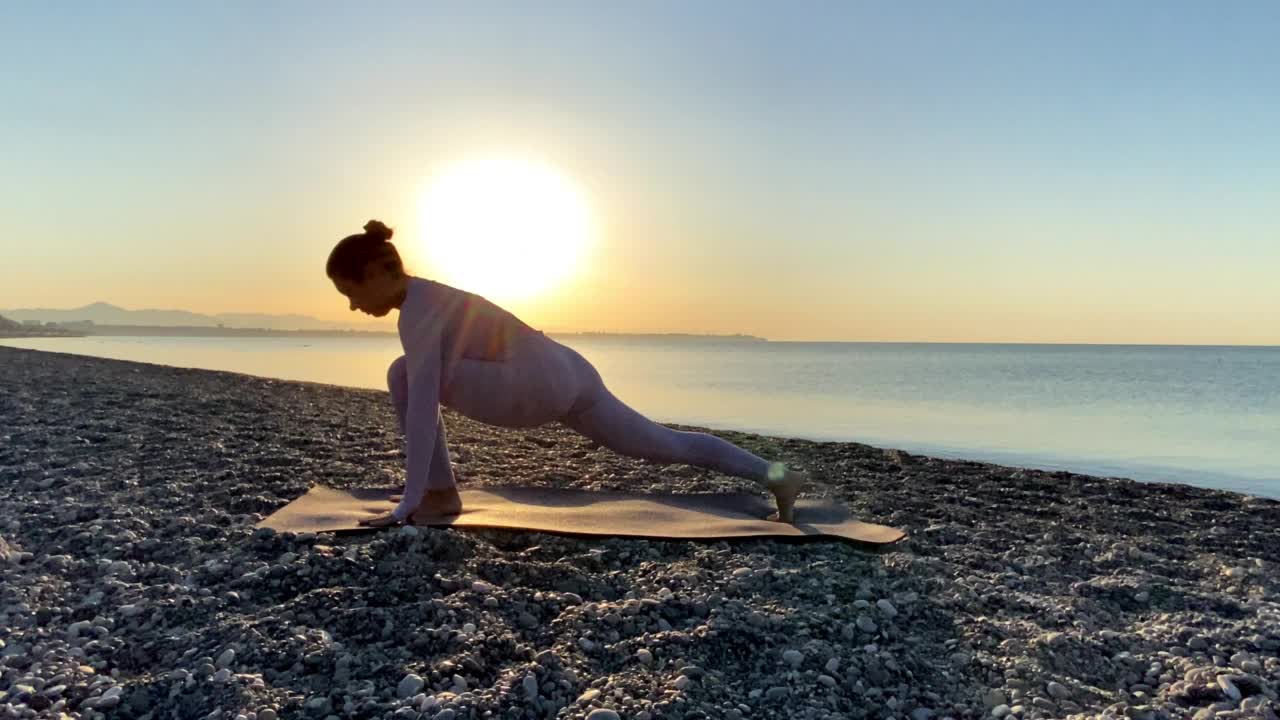 在日出或日落时，健康的年轻女子在平静的海面附近的海滩上练习瑜伽视频素材