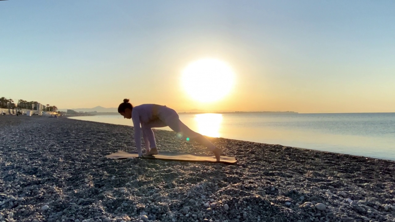 在日出或日落时，健康的年轻女子在平静的海面附近的海滩上练习瑜伽视频素材