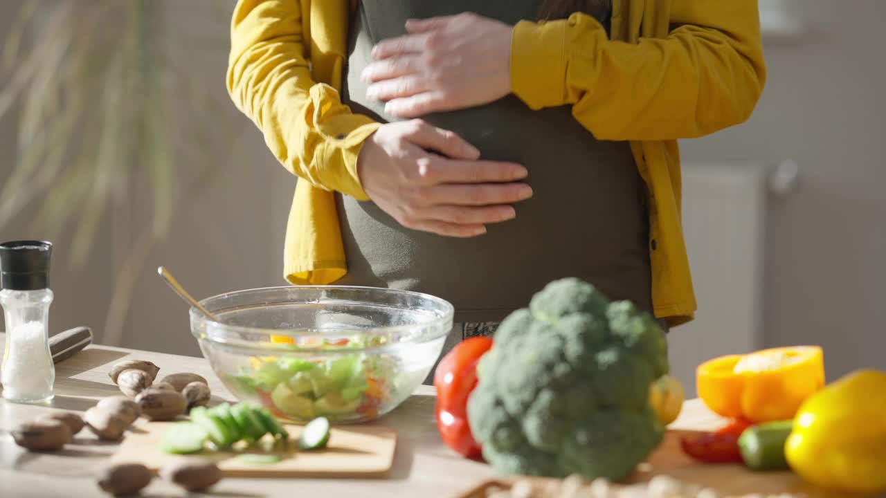 年轻的难以辨认的女人爱抚怀孕的肚子手势心形站在室内。快乐的白人期待烹饪健康的沙拉在家厨房室内享受休闲。视频素材