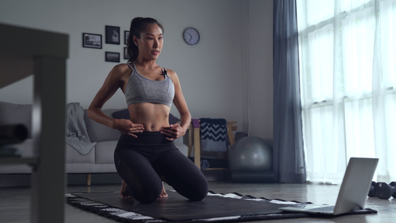 年轻的亚洲女性穿着运动服在家里锻炼，在网上观看健身视频或在线健身课程，使用笔记本电脑。视频素材