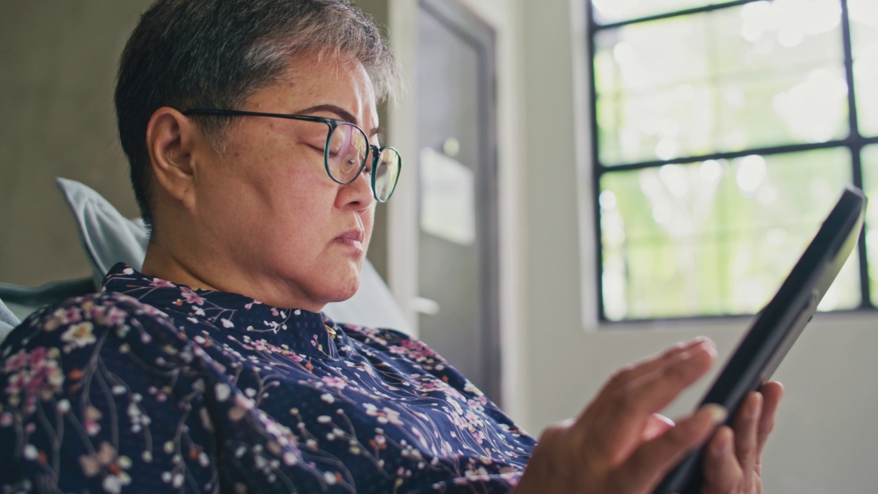 亚洲年长成熟女性在沙发上休息，独自在家使用数码平板电脑视频素材