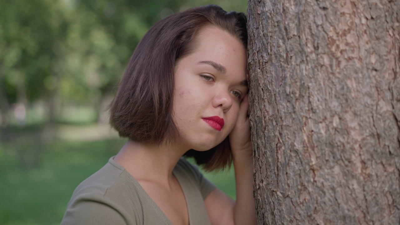 侧视图有思想的年轻女子与侏儒倚靠在树干上看向别处。迷人的白人小人儿在夏天的春天公园户外做梦。缓慢的运动。视频素材