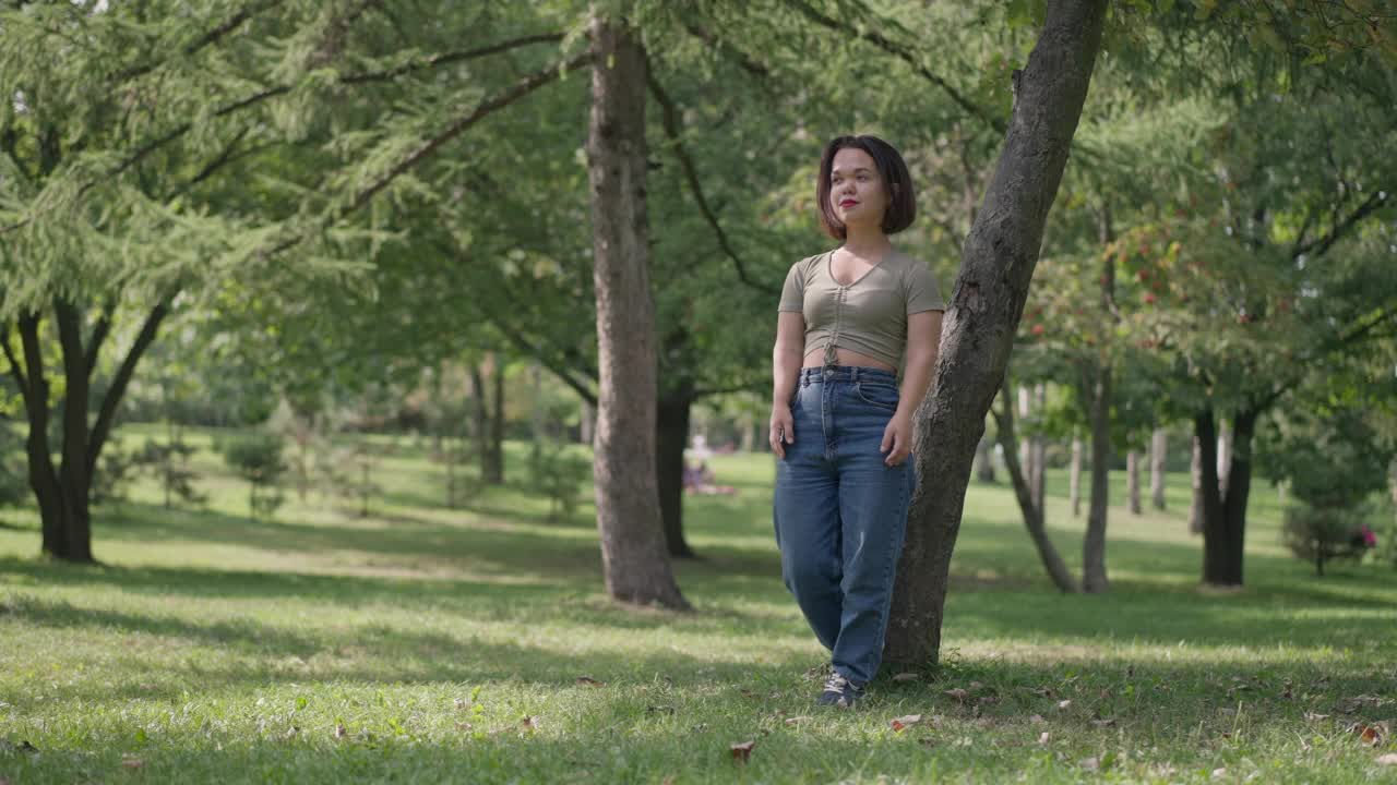 广角拍摄夏天公园森林在阳光灿烂的一天与白人年轻的小女人靠在树干望向远处站在右边。开心放松的侏儒症患者在户外做梦。视频素材