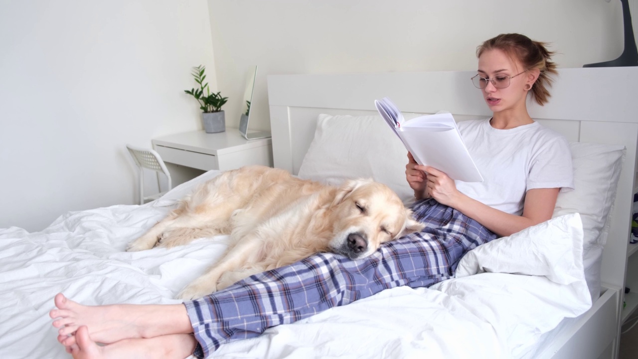 一个漂亮的女孩和她的金毛猎犬朋友在卧室里看书。一个年轻的女人和她的狗躺在床上。视频素材