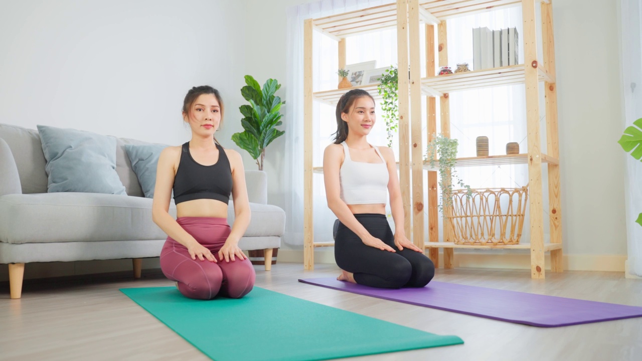 亚洲活跃的女性朋友在家里一起做瑜伽普拉提锻炼。漂亮的女孩穿着运动服，用垫子在家里的客厅里利用空闲时间进行保健健身。视频素材