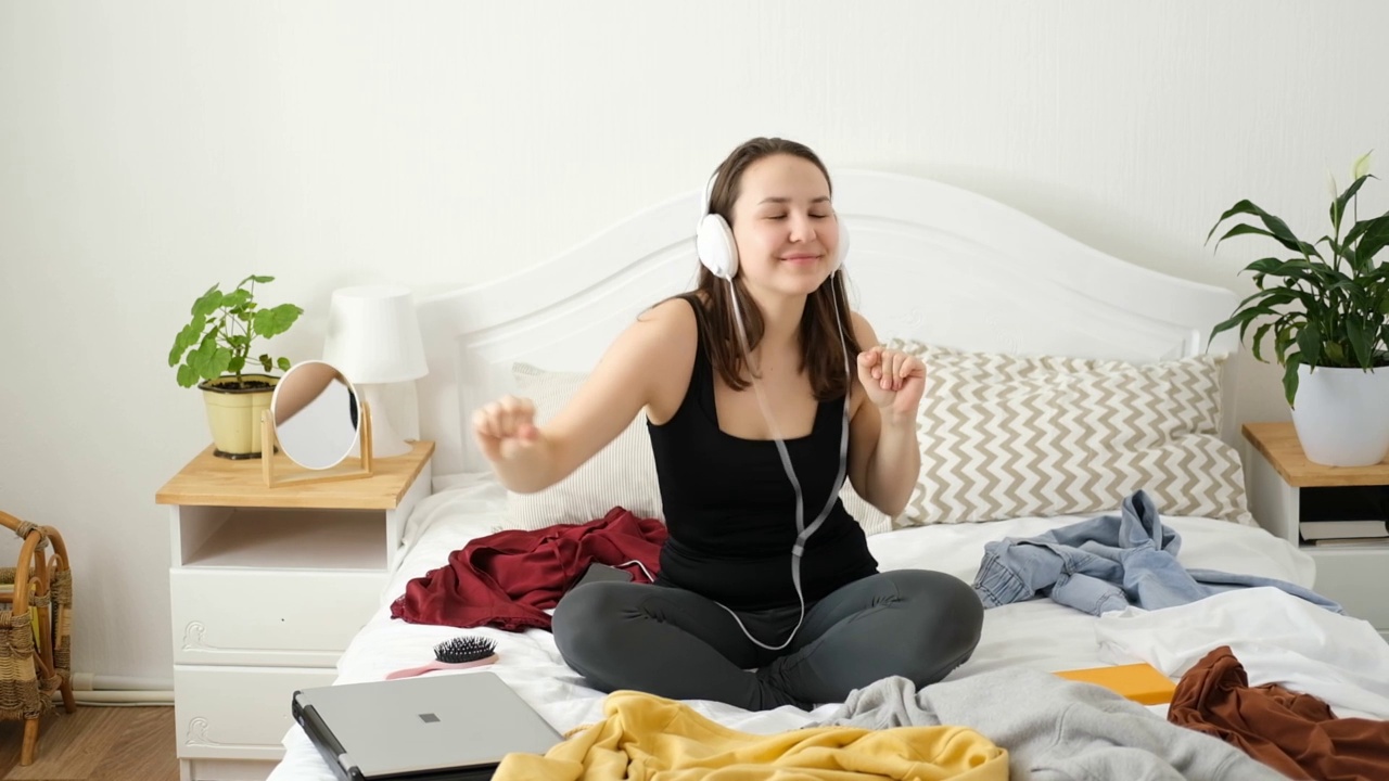 快乐的年轻白人黑发女子坐在床上戴着耳机，听音乐和跳舞，放松，抗压力。视频素材