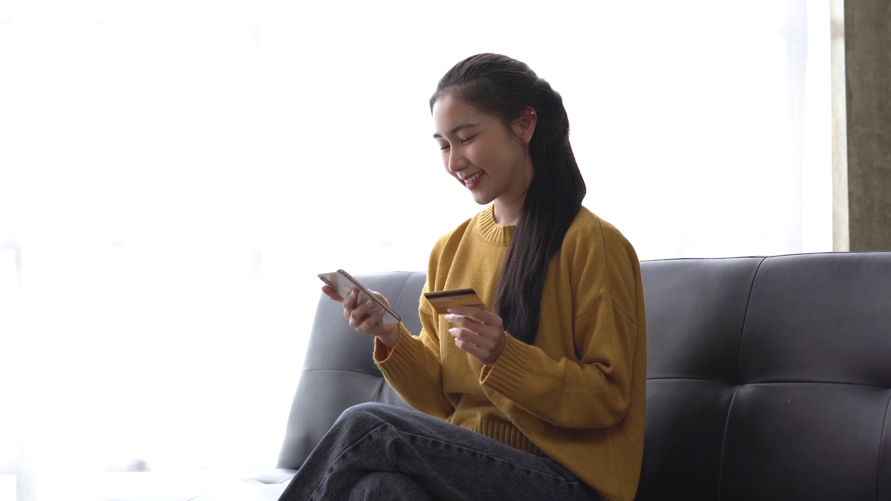 年轻的亚洲女性高兴地在沙发上用信用卡订购网上购物产品。视频素材