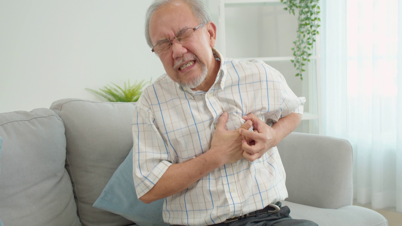 资深男性亚洲人遭受严重的疼痛在他的胸部心脏病发作坐在家里的沙发上。老人把他的手放在他的左胸口，因为他的心突然疼痛。心脏病。视频素材