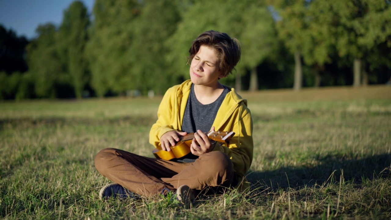 天才自信浪漫的少年坐在阳光灿烂的夏日草坪上弹奏尤克里里琴。熟练的白人青少年音乐家肖像享受户外爱好在夏季公园。视频素材