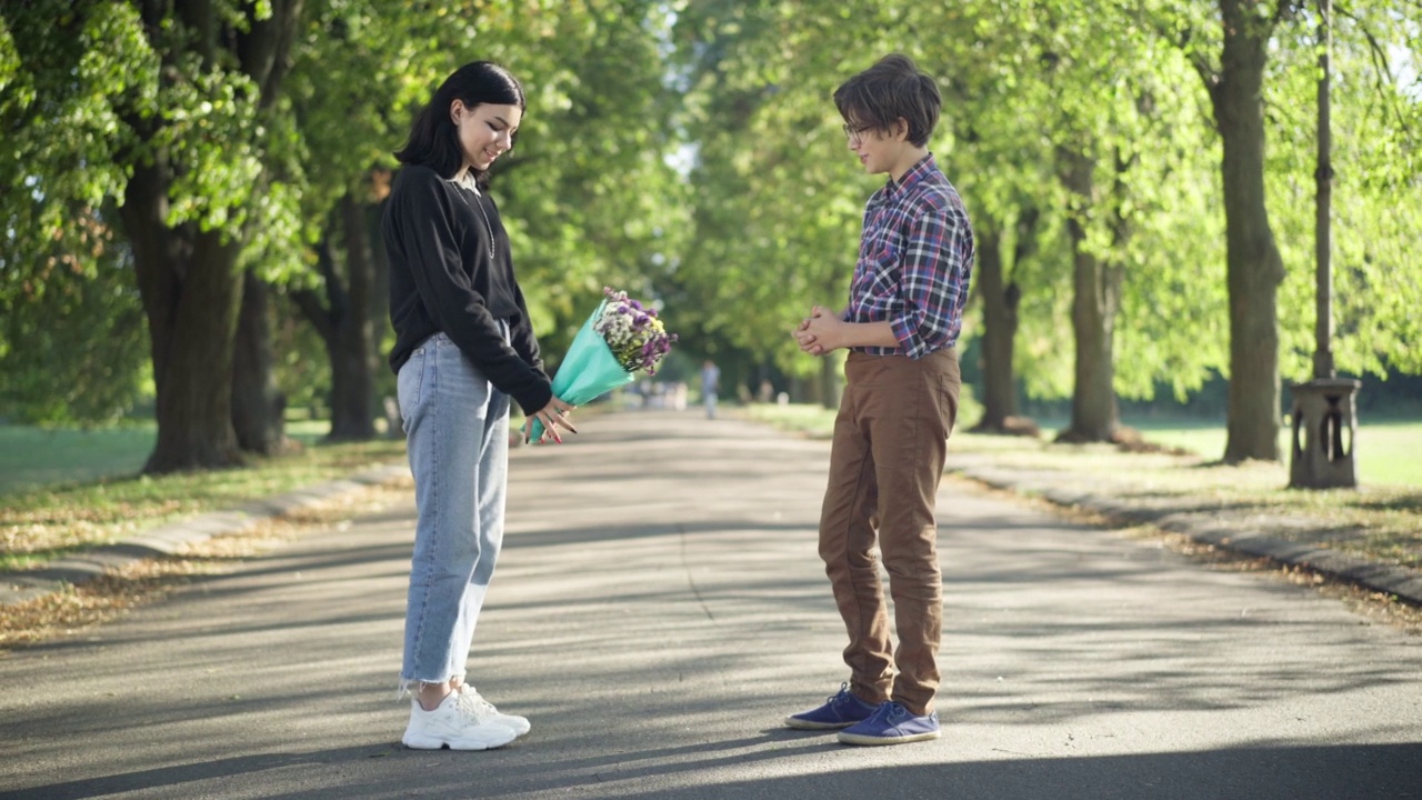 害羞的高加索青少年男孩和女孩约会在阳光明媚的夏季公园在周末。侧面的积极微笑的青少年夫妇谈论通过花束的广泛镜头。初恋的概念。视频素材