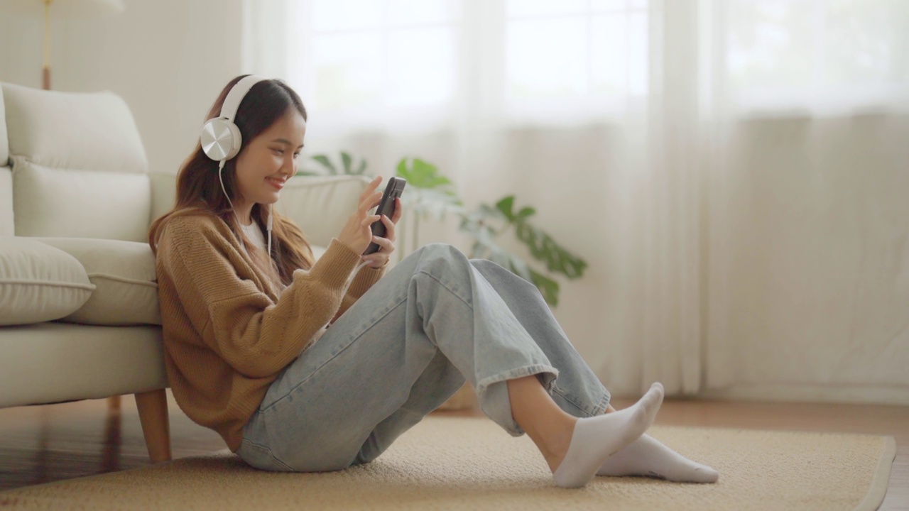 快乐的年轻亚洲女人在家里放松。女性微笑着坐在沙发上，手里拿着手机。女孩用视频打电话给朋友视频素材