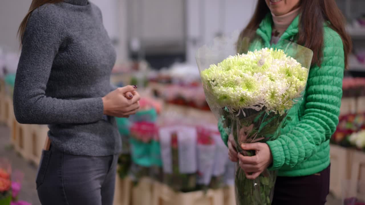 侧视图认不出来的女人通过一束美丽的鲜花站在室内。满意的买家和专业的卖家在花店洽谈买卖植物。视频素材