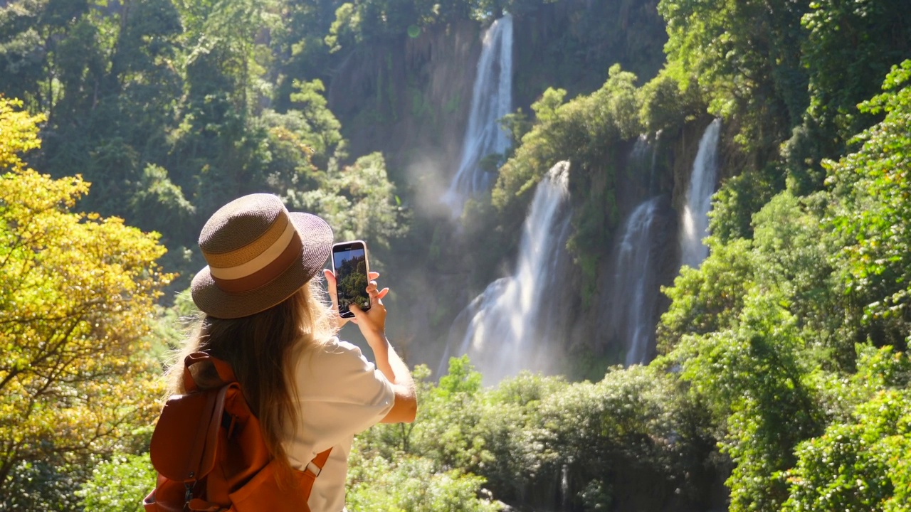 快乐的女性游客与背包旅行。漫游癖、旅游概念。年轻的女旅行者在她的智能手机相机上为社交媒体制作了令人惊叹的瀑布和热带自然的垂直视频视频素材