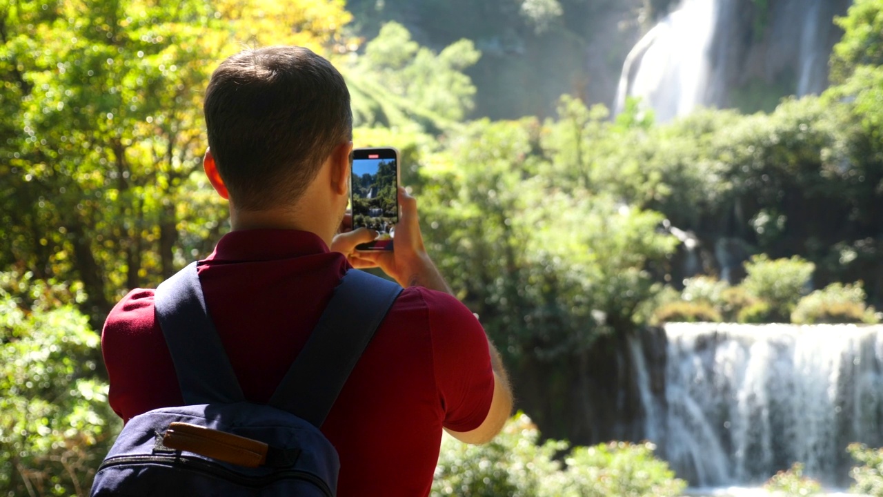 快乐的男性游客与背包旅行。漫游癖、旅游概念。年轻的旅行者在他的智能手机相机上为社交媒体制作了令人惊叹的瀑布和热带自然的垂直视频视频素材