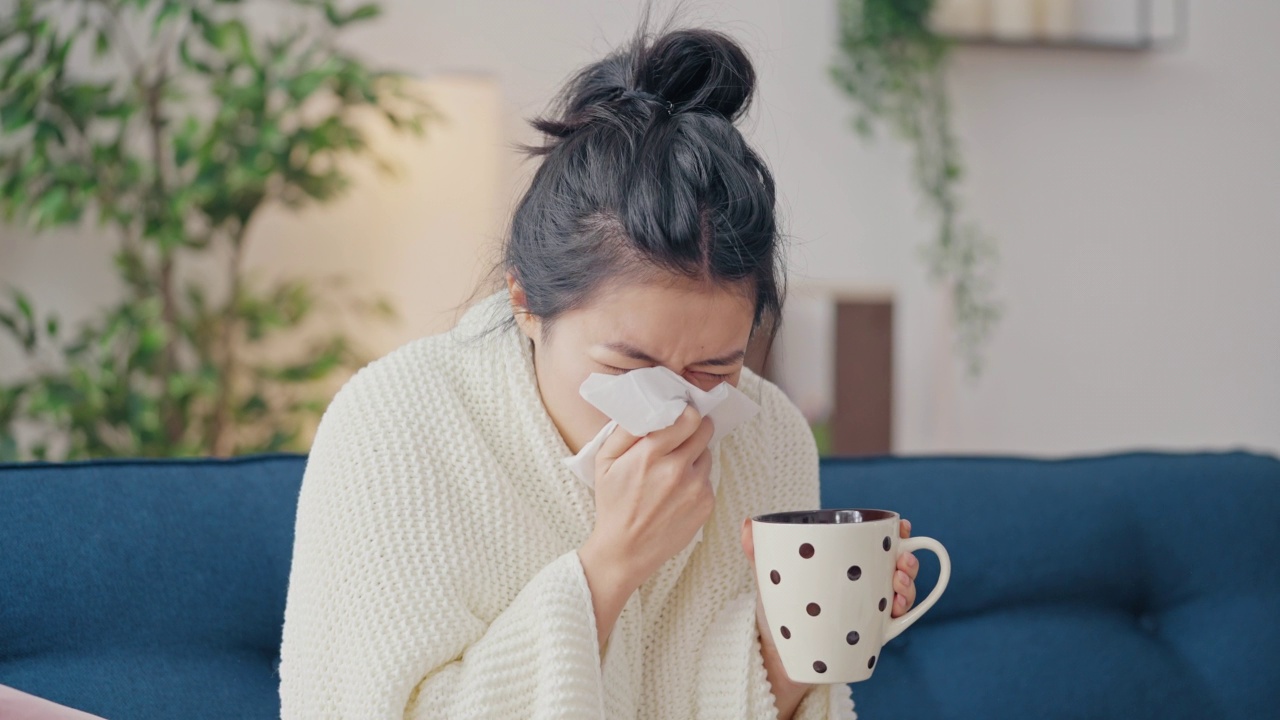 生病的亚洲妇女裹着毯子患流感在家，喝着热茶视频素材