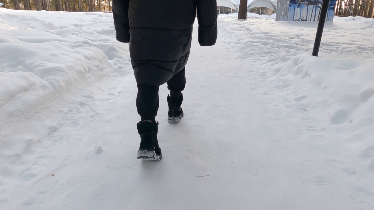 背影的女人腿在黑色的衣服和靴子走在冬天的雪路径在公园。的脚。特写镜头。后面的观点。裁剪。视频素材