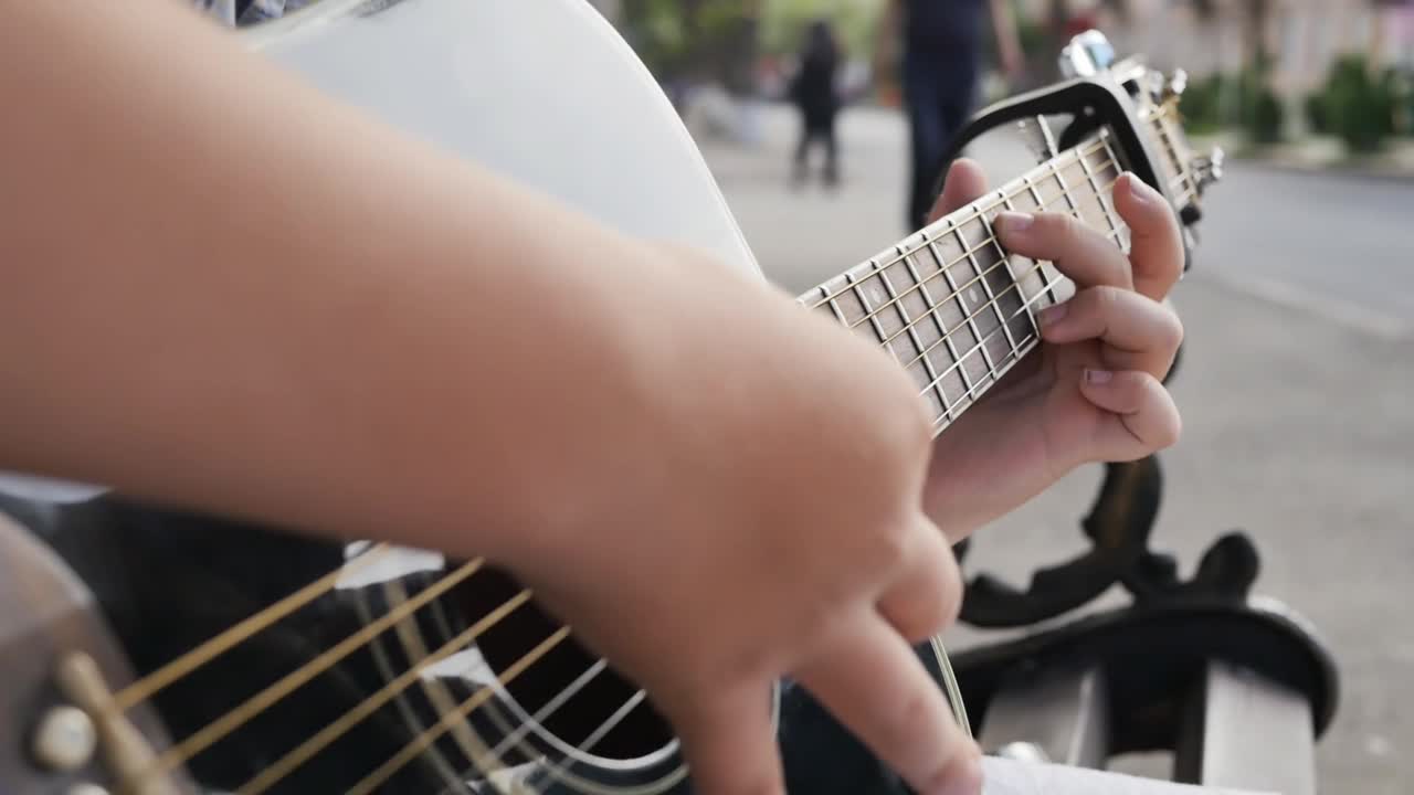 一个十几岁的男孩手里拿着一把原声吉他，晚上在户外的长凳上演奏。那个人的手指把琴弦捏成和弦，弹奏出一段安静的旋律视频素材