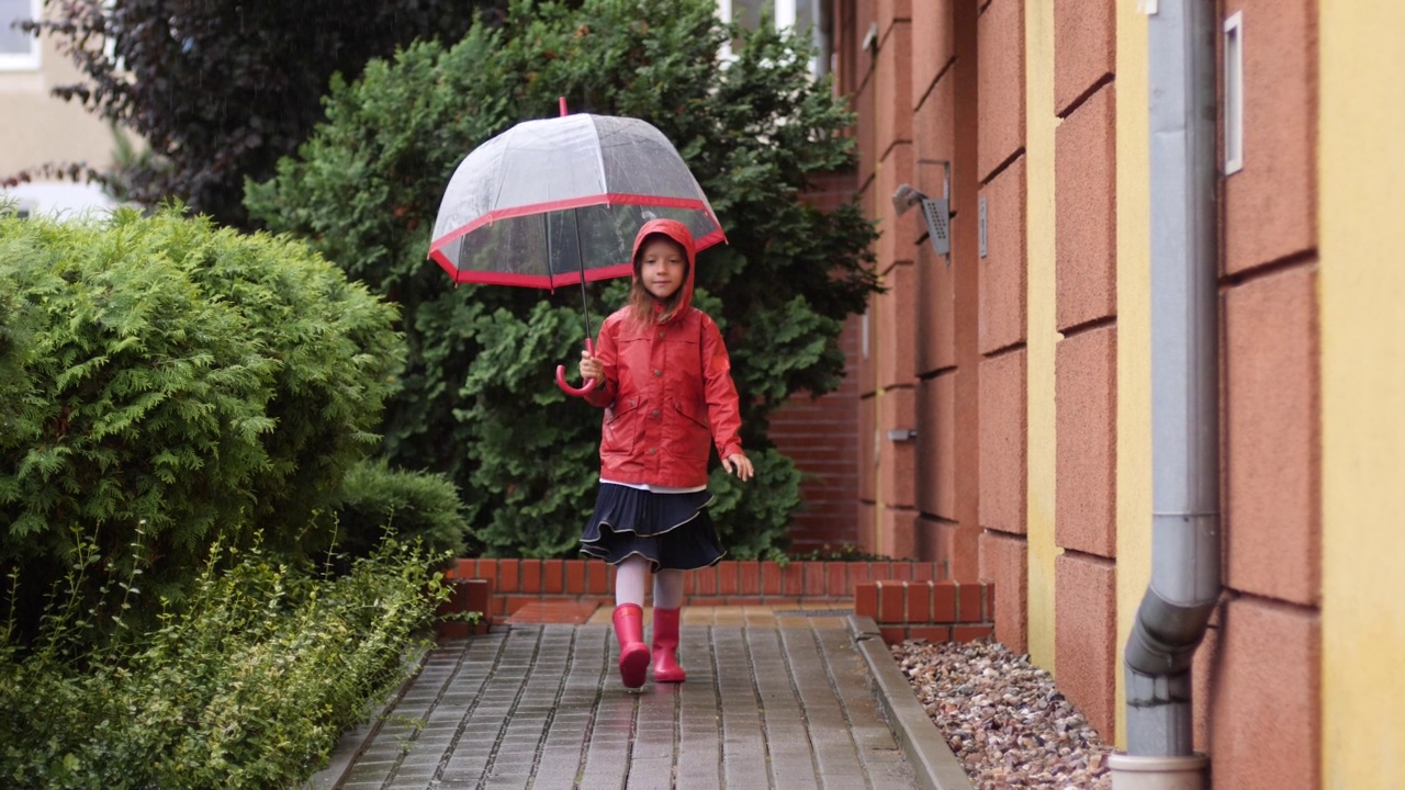 快乐的小女孩穿着红色雨衣和雨靴，撑着雨伞，喜欢在秋天的户外散步视频素材