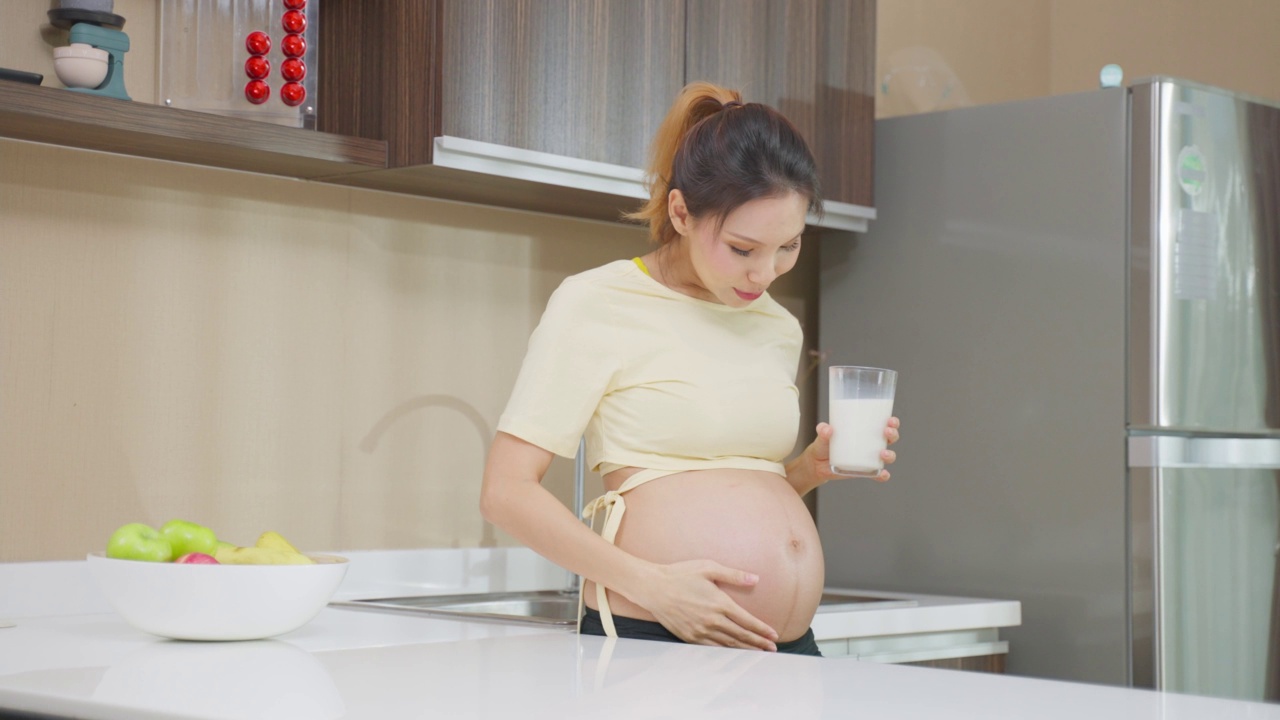 亚洲美女怀孕站在家里喝牛奶。迷人的孕妇站在子宫里抚摸宝宝，喝鲜奶健康饮料，在厨房里感受幸福。视频素材