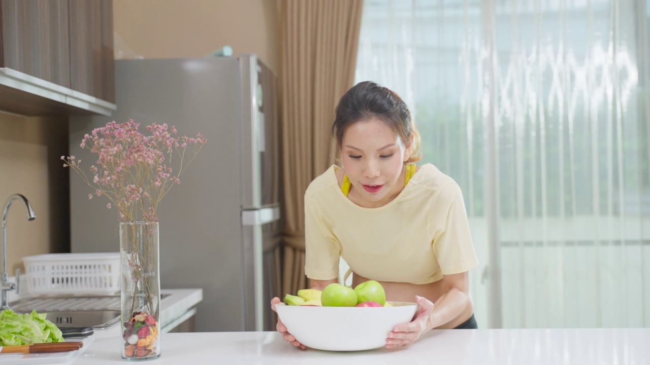 亚洲年轻孕妇的肖像在厨房的碗里拿着水果。迷人的怀孕女孩站在室内感觉快乐，享受吃健康的食物，然后看着镜头。视频素材