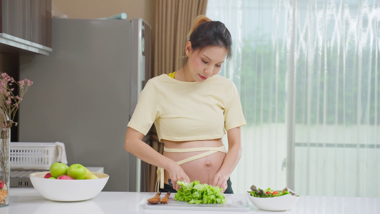 亚洲美丽的孕妇在家里厨房烹饪健康的食物。迷人的怀孕女孩站在厨房的室内感到快乐，享受在碗绿色蔬菜沙拉的健康在家里视频素材