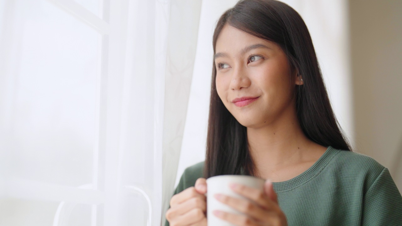 年轻的亚洲女子站在窗边，拿着杯子在家里的卧室，她早上醒来后喝牛奶视频素材