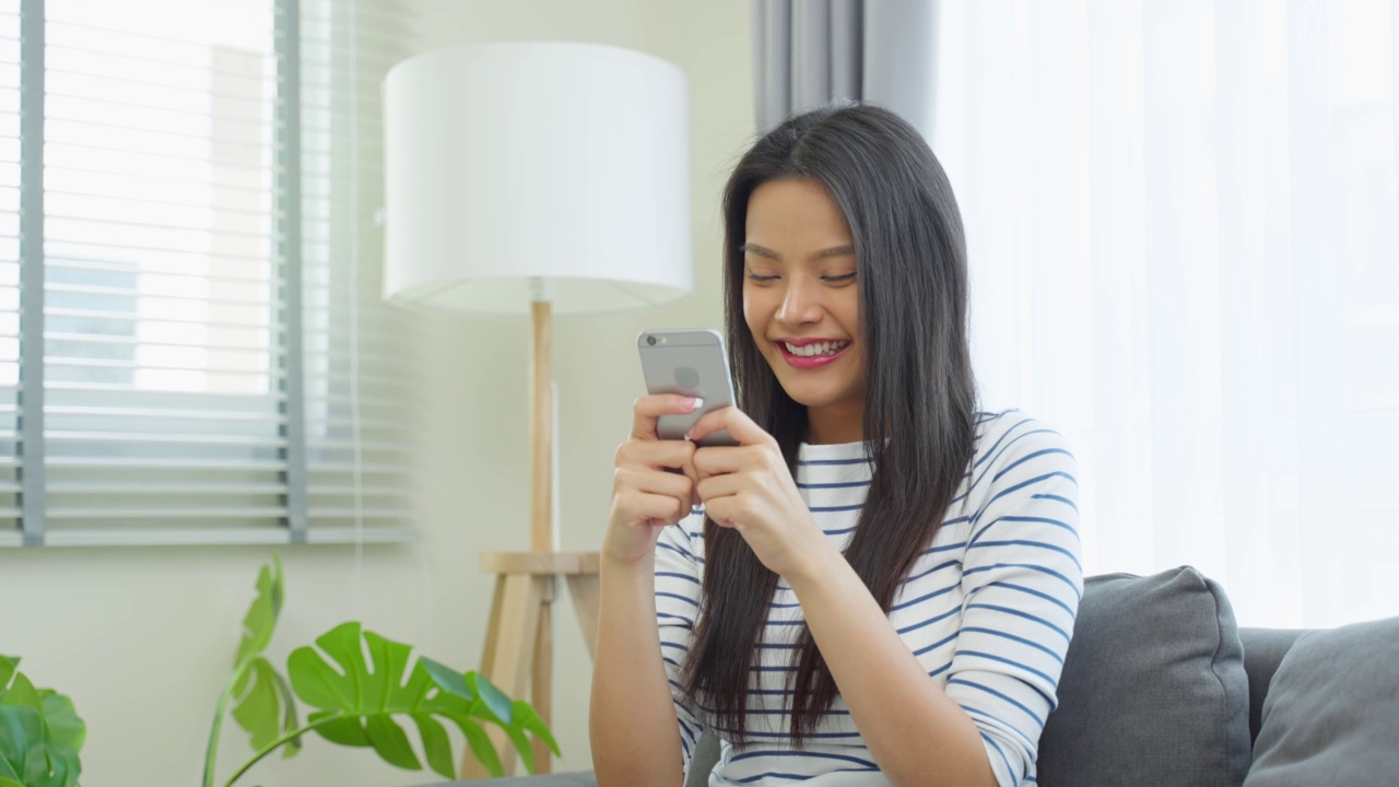 亚洲美女坐在家里的沙发上用手机聊天。快乐迷人的年轻女孩在家里度过休闲时间，感到放松，享受在客厅用智能手机交流和讨论。视频素材