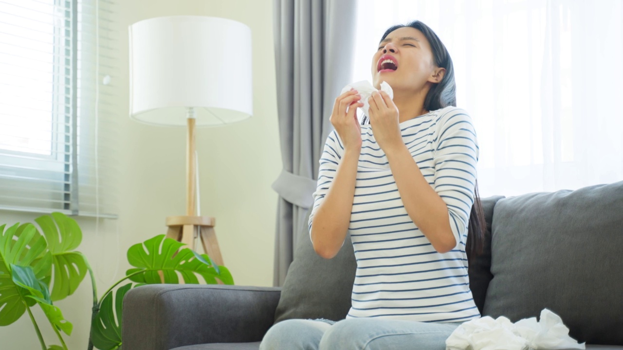 亚洲美女在客厅流鼻涕和打喷嚏。有魅力的年轻女性坐在沙发上感觉不好，患上了过敏，然后在家里打喷嚏时用纸巾捂住鼻子。视频素材