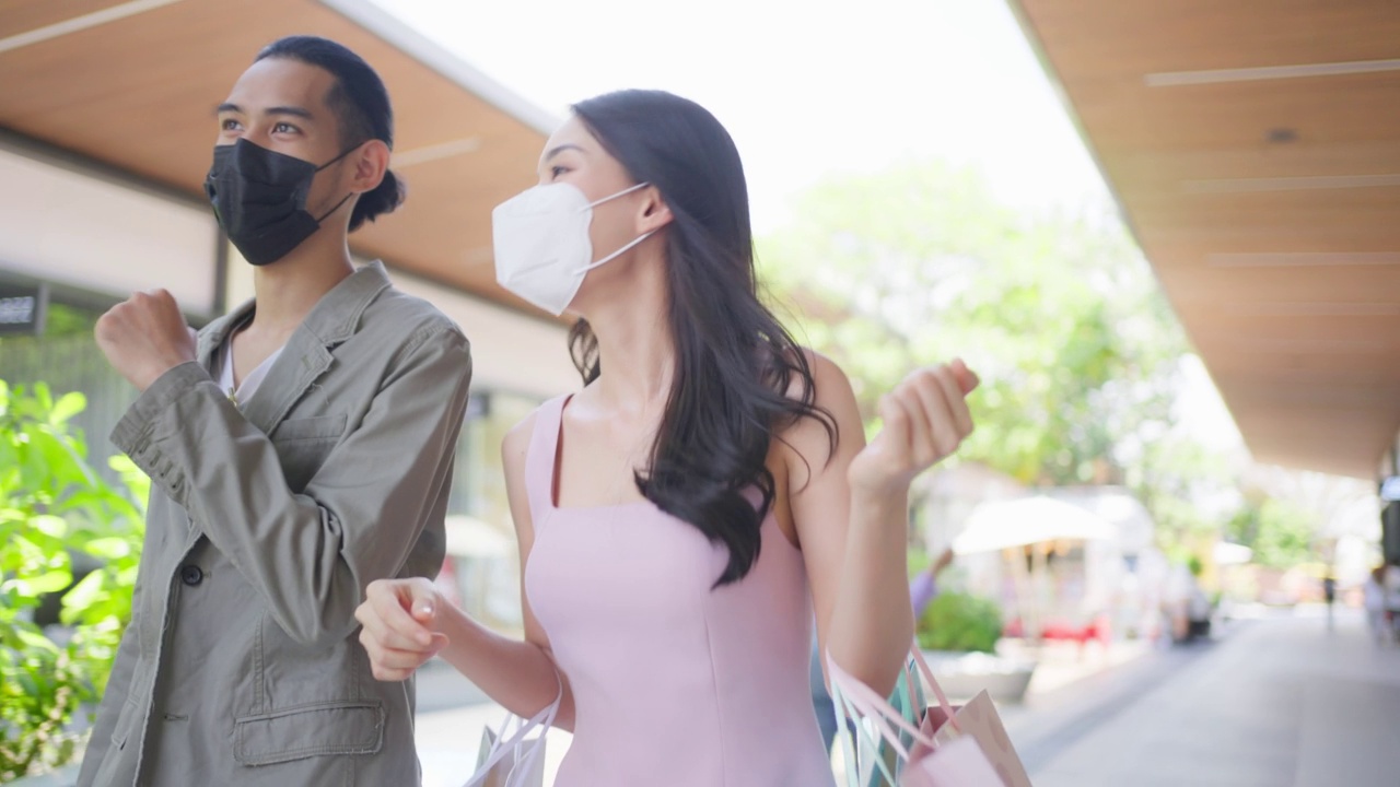 亚洲青年男女在百货公司户外购物。迷人的情侣戴上口罩，拎着购物袋，幸福地走在一起，在商场购物。视频素材