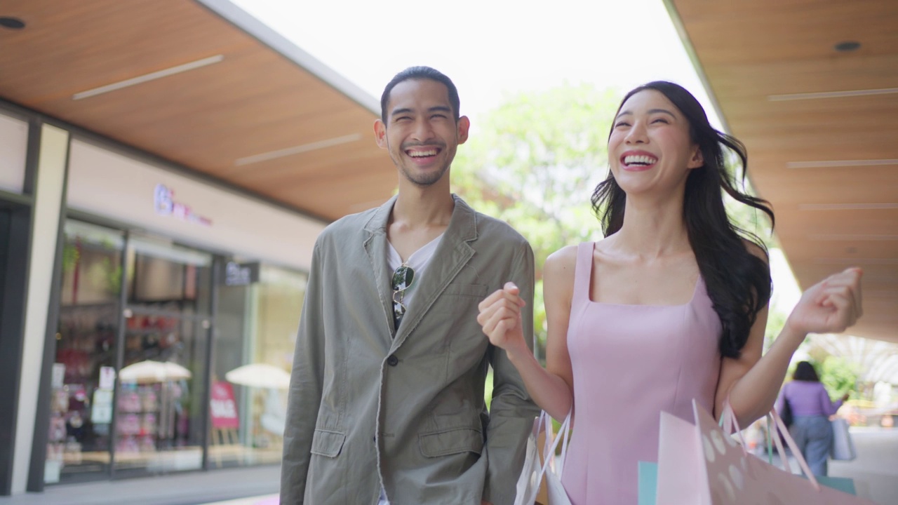 亚洲青年男女在百货公司户外购物。迷人的情侣拿着购物袋，然后幸福地走在一起，享受购物中心购物。视频素材