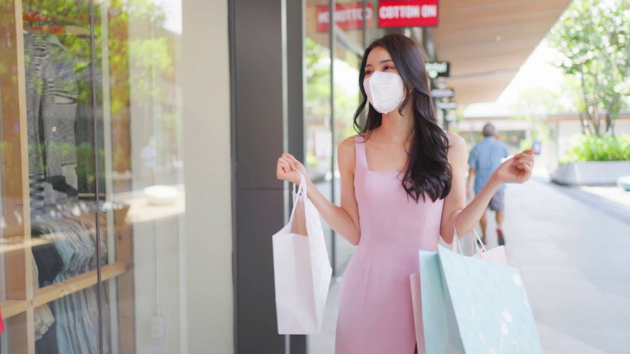 亚洲美丽的年轻女子在百货公司户外购物。迷人的女性戴着口罩，拎着购物袋，在购物中心快乐地独自行走。视频素材