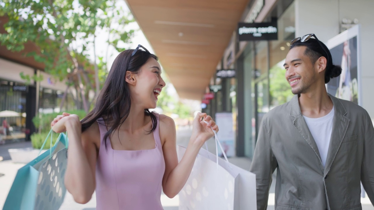 亚洲青年男女在百货公司户外购物。迷人的情侣拿着购物袋，然后幸福地走在一起，享受购物中心购物。视频素材