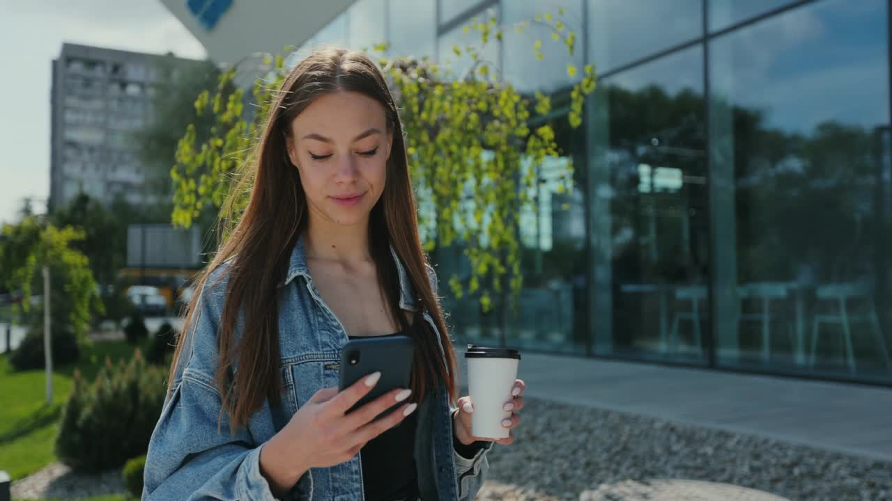 拿着智能手机和咖啡杯的女孩。美女在街上用手机喝外卖咖啡。视频素材