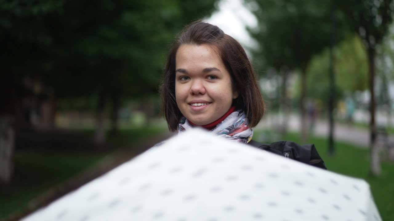 一个积极微笑的小人儿站在雨天的绿色城市街道上看着相机。快乐自信的白人小女人摆出的慢动作户外旋转伞。视频素材