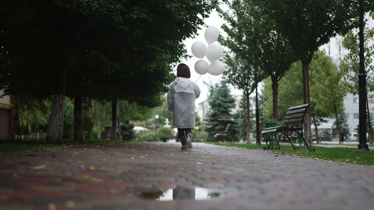 宽镜头的背影，年轻的小女人在雨衣与白色气球走在户外慢动作。轻松自信的白人小人儿漫步在城市的小巷里。视频素材