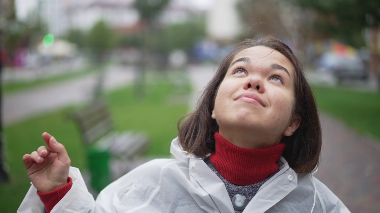 黑发美女脱下雨衣兜帽，抬头微笑。幸福自信的白人小人物肖像在雨天欣赏云在城市。视频素材