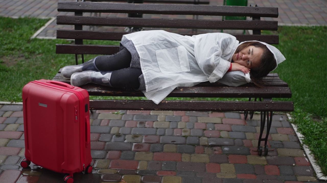 广角镜头悲伤无望的小女人穿着雨衣躺在户外的长凳上，旅行袋站在一边。沮丧悲伤的白人小人物的肖像与行李在城市街道上。视频素材