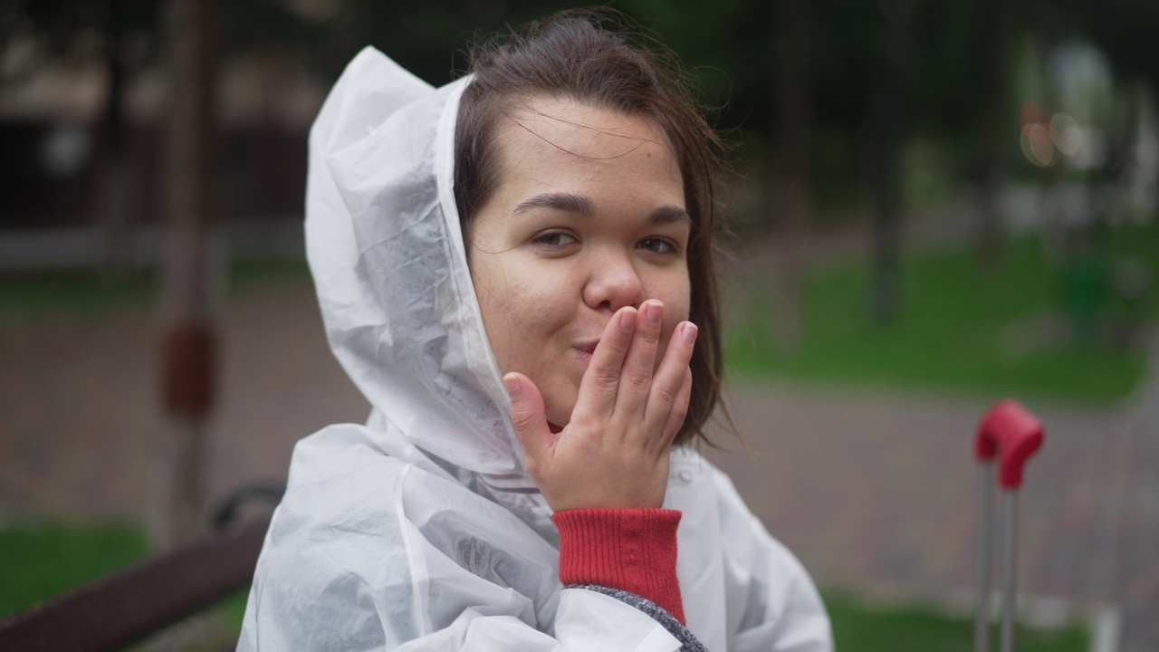 积极微笑的小女人在雨衣手势飞吻看着镜头。特写肖像快乐自信的白人小人物摆出的慢动作在阴雨天户外。视频素材