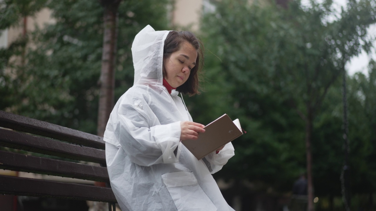 悠闲的小女人穿着雨衣坐在长椅上看书。在阴雨天的户外公园里全神贯注的白人小人物的肖像。缓慢的运动。视频素材