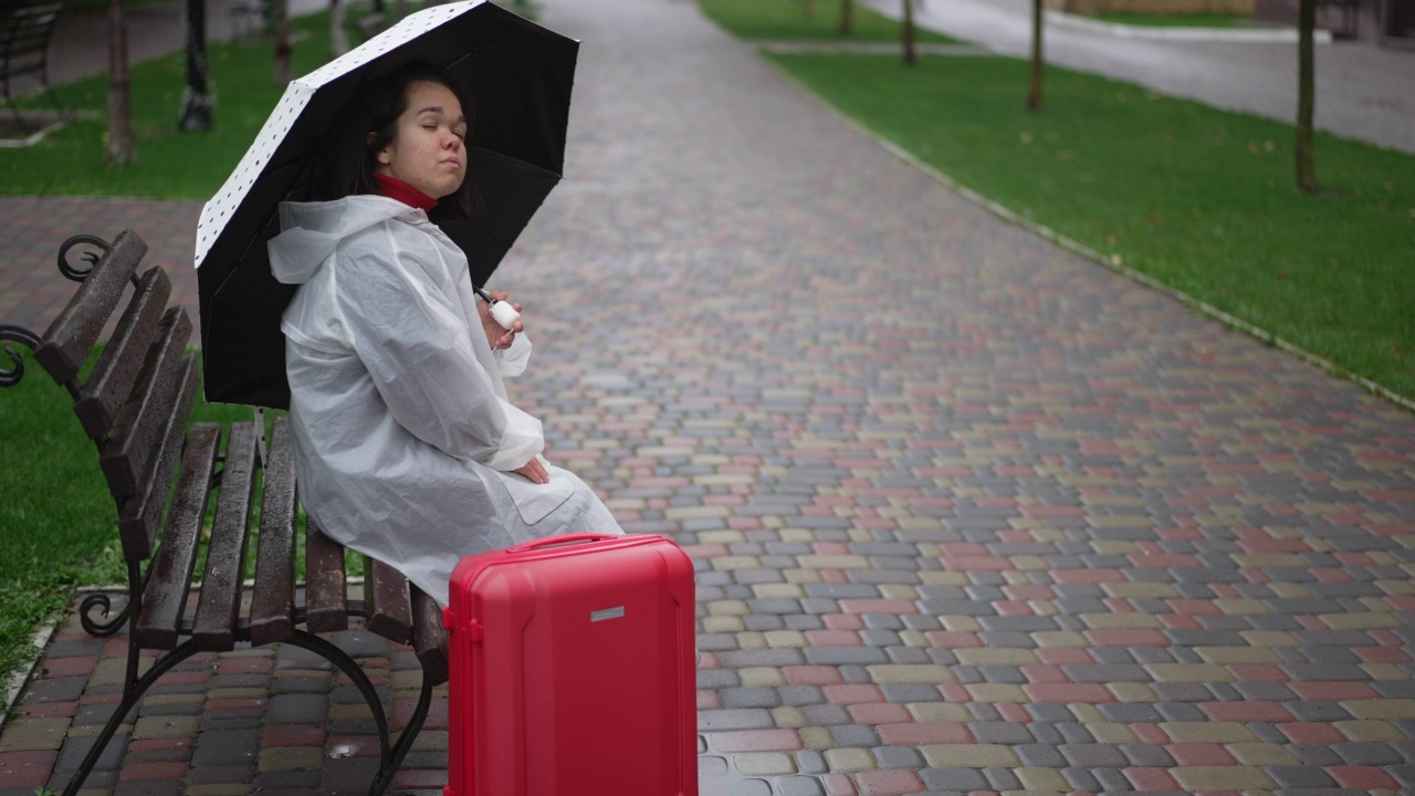 忧伤孤独的小女人穿着雨衣，拿着雨伞和行李箱坐在长椅上思考。侧视图广角年轻的高加索小人儿独自旅行在城市户外。缓慢的运动。视频素材