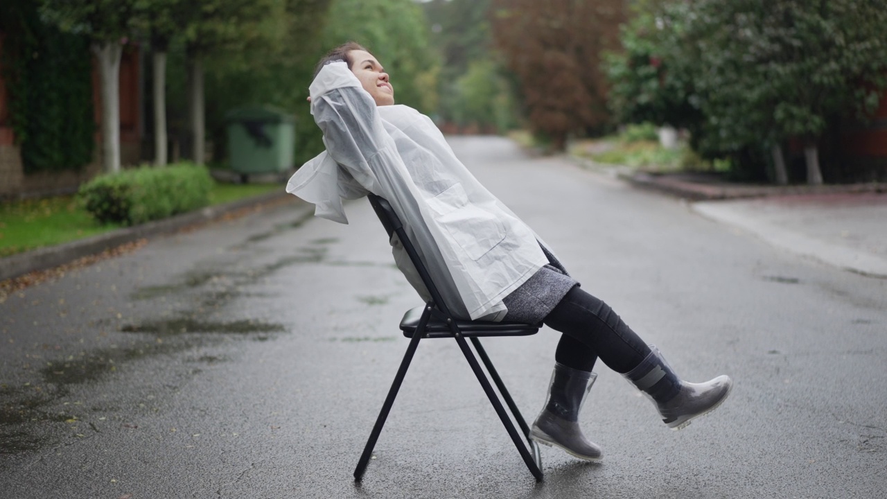 悠闲的小人儿穿着雨衣和胶靴坐在椅子上微笑着享受郊区阴天的雨天。侧视图广角自信的高加索小女人休息在道路户外。视频素材