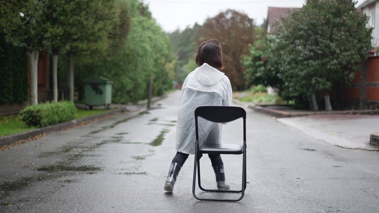 后视图小女人穿着雨衣坐在郊区道路中间的椅子上。轻松悠闲的白种人积极的小人儿穿着雨衣在户外欣赏雨天。视频素材