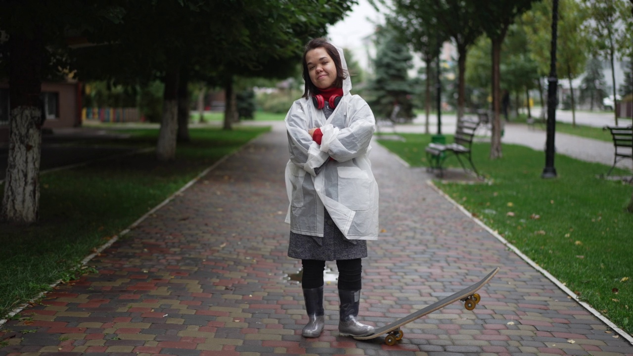 自信的小女人穿着雨衣站在户外滑板交叉双手看着相机的肖像。微笑积极的白人小个子在城市街道上享受运动和休闲。视频素材