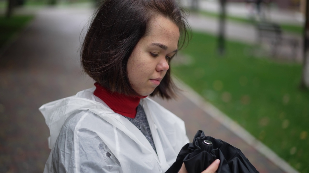 侧面视图特写自信的小女人折叠伞在慢动作站在户外。美丽的白种人小个子在阴天的城市街道上穿着雨衣。视频素材