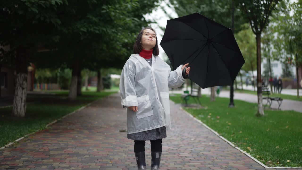 宽镜头年轻美丽的小女人在雨衣和胶靴思考仰望站在户外的雨伞。白种人小人儿的肖像在城市街道上休息。视频素材