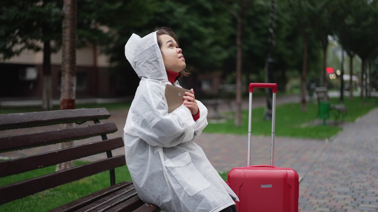 年轻的小个子抱着书，抱着行李箱坐在长凳上。穿着雨衣的白人小妇人在城市户外享受业余旅行的肖像。缓慢的运动。视频素材